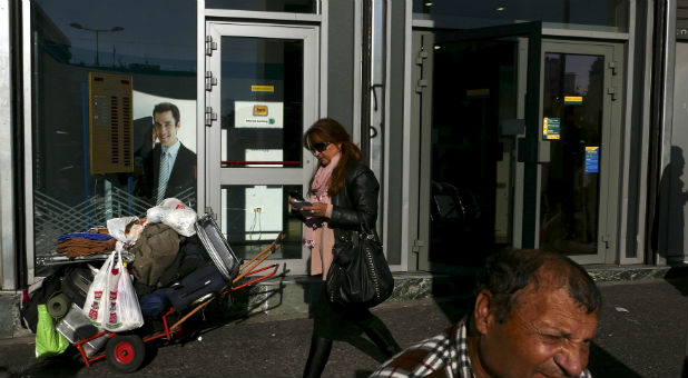 A woman exits a National Bank branch as a man sits nearby in Athens, Greece,