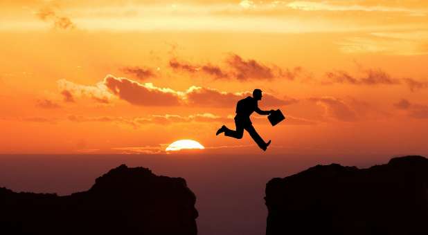 Man holding briefcase leaping from one cliff to another.